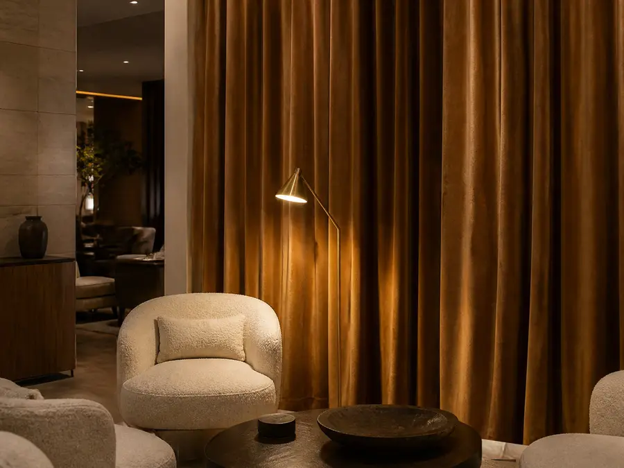 Hotel lounge in warm light, camel velvet curtains with deep folds, a brass reading lamp and textured cream armchairs